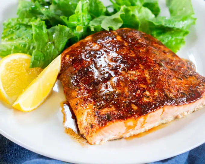 A close up of a baked filet of Salmon drizzled with brown butter and lemon juice sitting on a white plate. A lettuce salad and Lemon slices sit in the background.