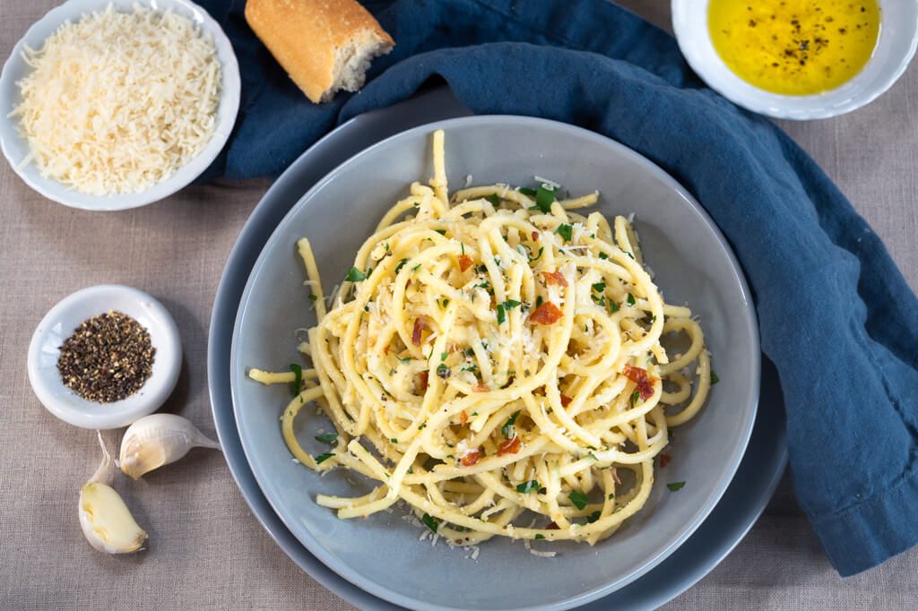 Top-down view of Bucatini pasta tossed with grated cheese, parsley, black pepper, and prosciutto in a gray bowl over a blue napkin. The same ingredients surround the dish in small white bowls.