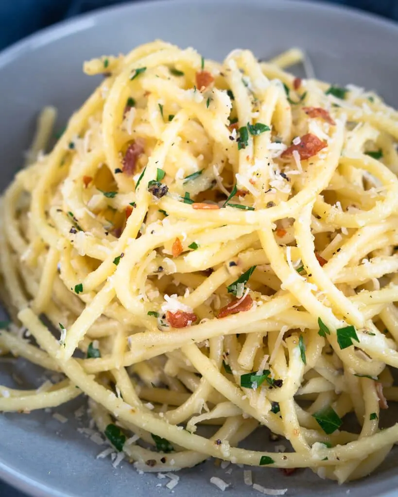 Front view of Bucatini pasta tossed with grated cheese, parsley, black pepper, and prosciutto in a gray bowl.