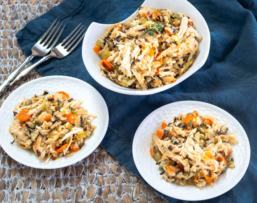 Top-down view of a white bowl heaping with chicken and rice filled with carrots, onions, and herbs over a blue cloth. Two white plates are in the foreground next to 2 forks.