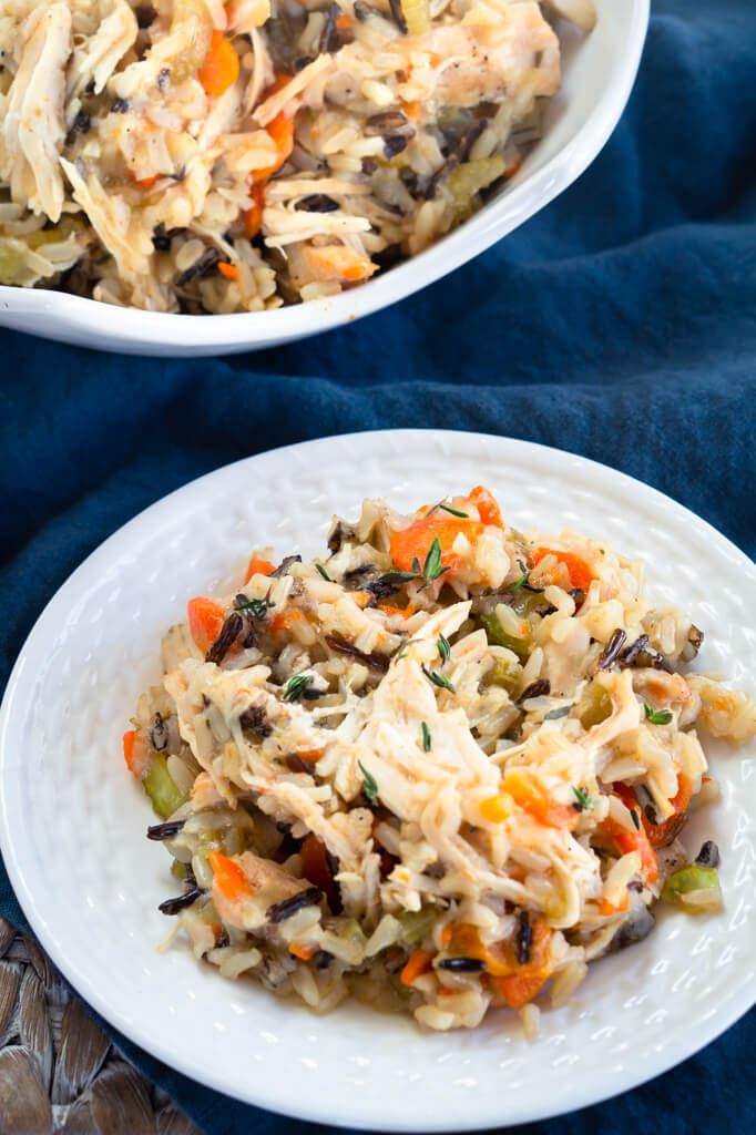 A white plate of chicken and rice filled with carrots, onions, and herbs over a blue cloth. A platter of the pressure cooker chicken and rice is in the background. Hostess At Heart
