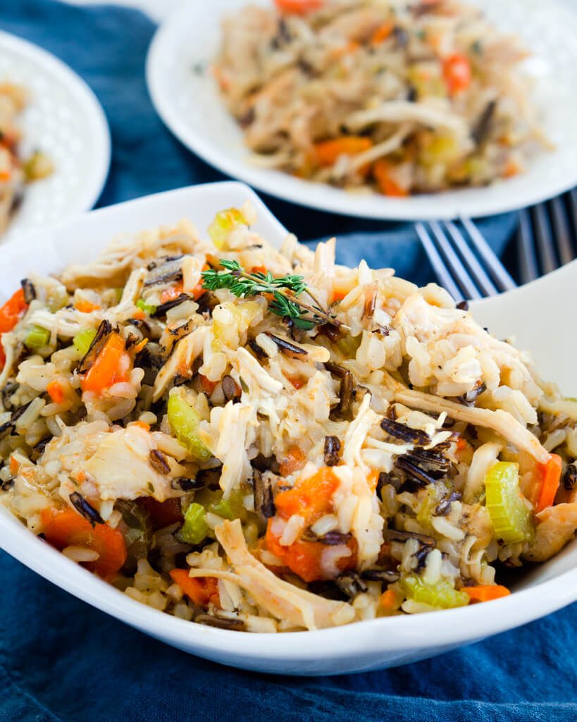 A white bowl  heaping with chicken and rice filled with carrots, onions, and herbs over a blue cloth. Two white plates are in the background. 