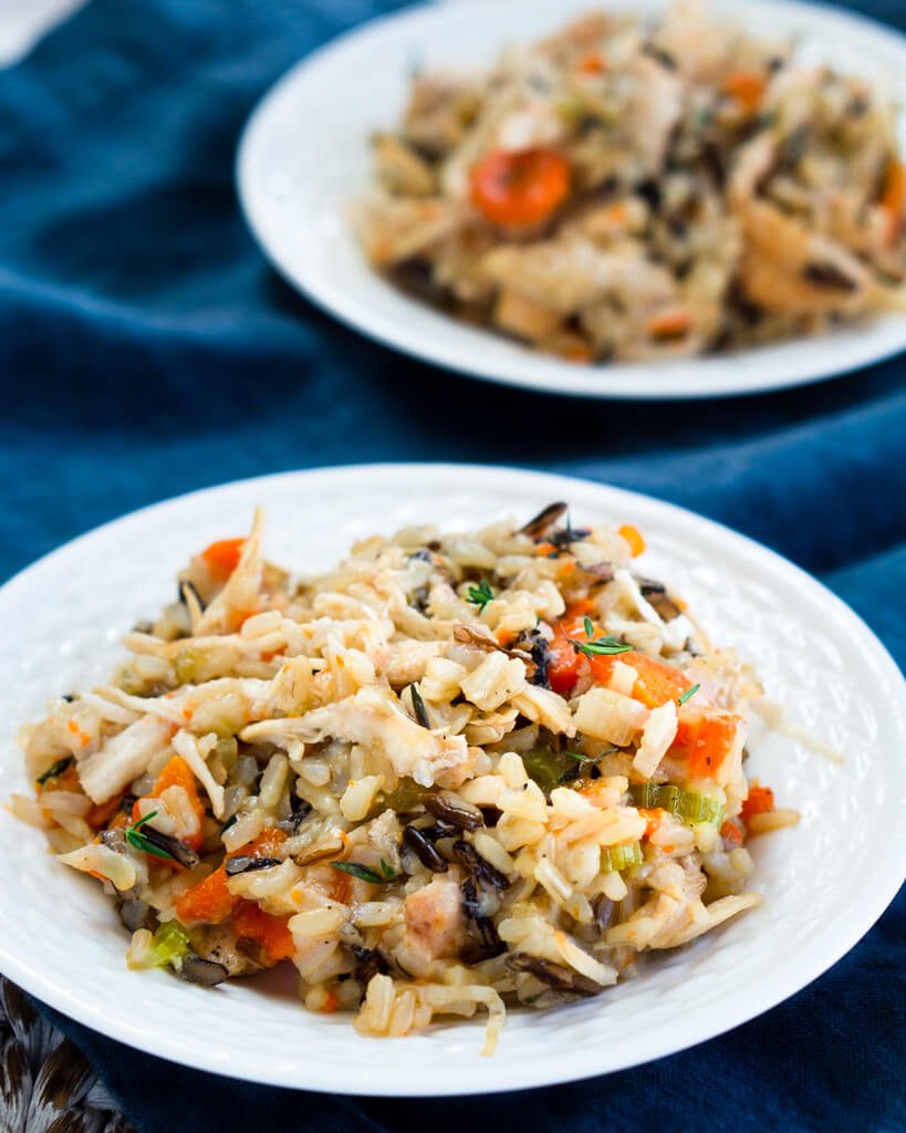 A white plate of chicken and rice filled with carrots, onions, and herbs over a blue cloth. A second plate is in the background.
