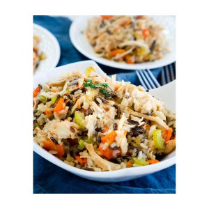 A white bowl heaping with one-pot chicken and rice filled with carrots, onions, and herbs over a blue cloth. Two white plates are in the background. Hostess At Heart