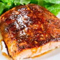 A close up of a baked filet of Salmon drizzled with brown butter and lemon juice sitting on a white plate. A lettuce salad and Lemon slices sit in the background.