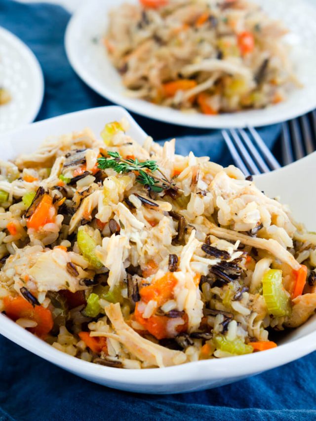 A white bowl heaping with chicken and rice filled with carrots, onions, and herbs over a blue cloth. Two white plates are in the background.