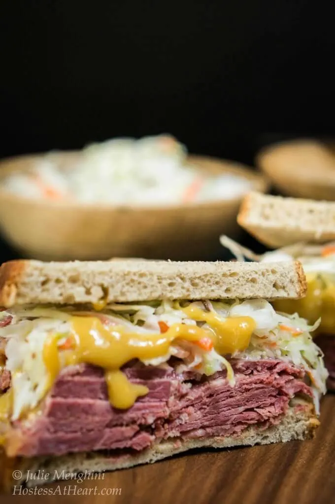 Stacked two halves of a corned beef sandwich with layers of brown sugar mustard and caraway slaw between to slices of rye bread.   A bowl of slaw sits in the background.