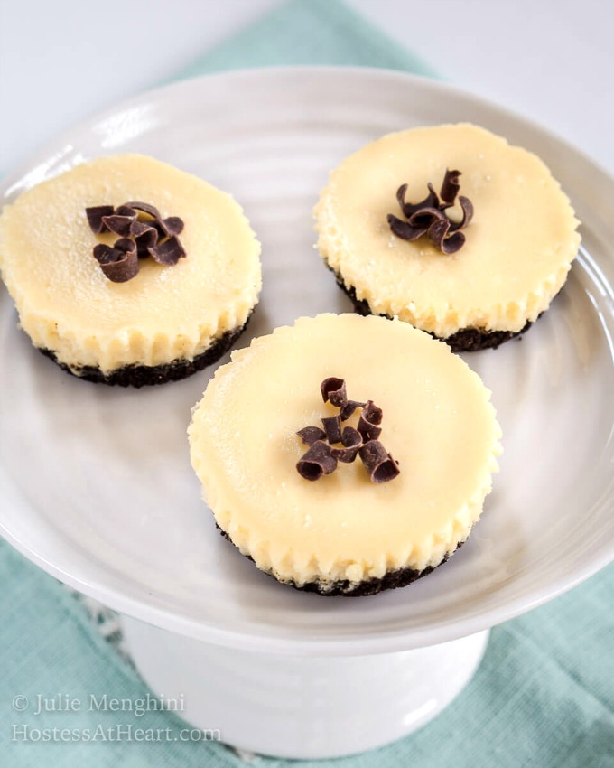 Top-down view of 3 Mini Cheesecakes have an Oreo crust, white cheesecake filling and topped with chocolate shavings. They sit on a small cake plate over a blue napkin. 