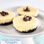 Mini Cheesecakes have an Oreo crust, white cheesecake filling and topped with chocolate shavings. They sit on a small cake plate over a blue napkin. The title "Chocolate Caramel Baileys Mini Cheesecakes appears in the bottom left corner.