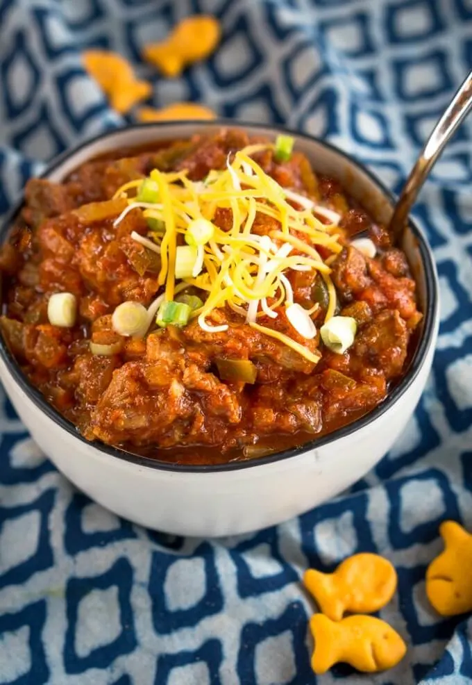 Top view of a white bowl with a black rim is filled with chili garnished with cheddar cheese and diced green onion. The bowl sits on a blue checked napkin and goldfish are scattered around the bowl.