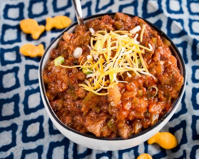 Top-down view of a white bowl with a black rim is filled with chili garnished with cheddar cheese and diced green onion. The bowl sits on a blue checked napkin and goldfish are scattered around the bowl.