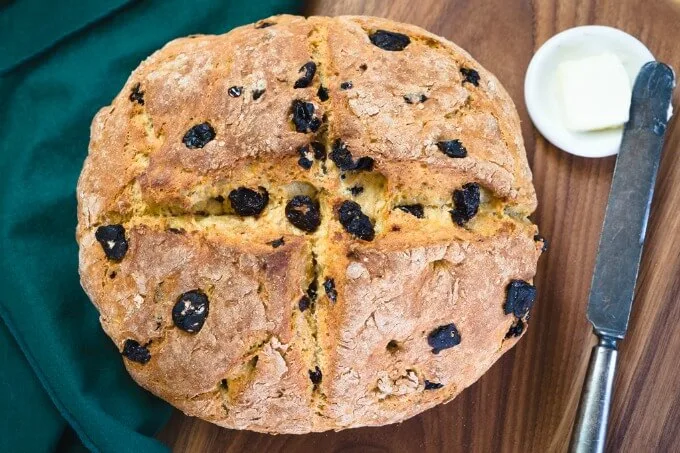 Top-down view of a whole loaf of Soda Bread dotted with cherries over a green napkin on a wooden cutting board.