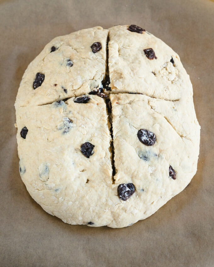 Unbaked top-down view of a whole loaf of Soda Bread dotted with cherries with a cross scored through the top on a piece of parchment paper. 
