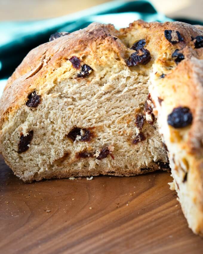 Front view of a loaf of Soda Bread dotted with cherries with a wedge removed next to a green napkin on a wooden cutting board.