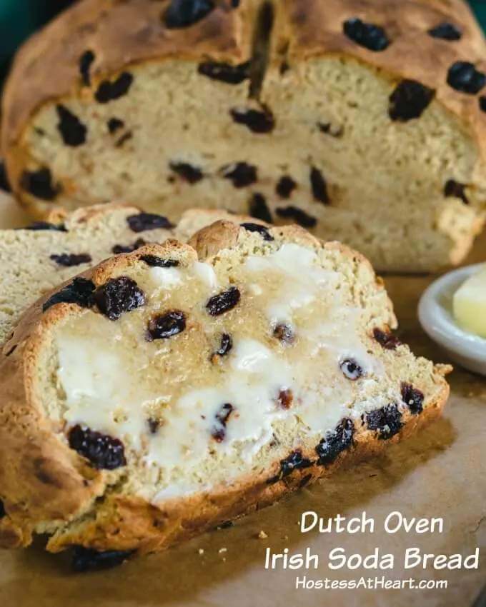 Level view of slices of soda bread dotted with raisins sitting on a wooden cutting bread. Butter is melting down the slice and a white bowl of butter sits to the side.