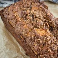 A loaf of Orange Pecan Streusel Coffee Cake with a thick brown sugar pecan streusel sitting at an angle on a piece of parchment paper.