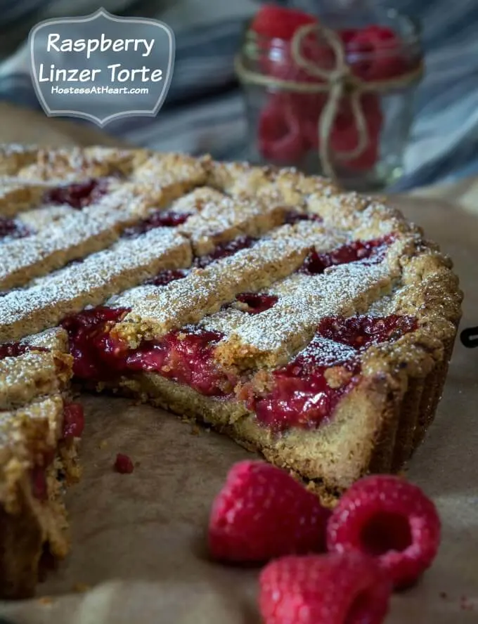 A tart with red raspberry filling and a lattice top dusted with powdered sugar with a piece missing Sitting on a piece of parchment paper. Fresh raspberries sit in the front and in a jar in the back.