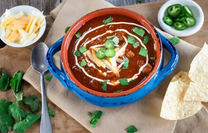 Top-down view of a blue bowl of Chili Con Carne garnished with cheese, jalapenos, and cilantro. A spoon sits to the side over a linen napkin.