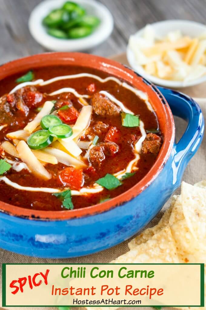 Angled view of a blue bowl of Chili con carne garnished with cheese, jalapenos and a swirl of sour cream. Tortilla chips and a white bowl of cheese sits in the background.