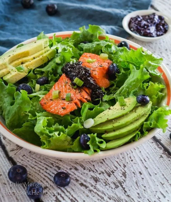 A white bowl with an orange rim is filled with green lettuce that\'s adorned with a thick piece of salmon topped with a balsamic blueberry sauce. Sliced avocado sits on each side of the salad and fresh blueberries are mixed into the greens over a white board.