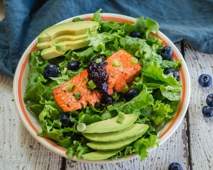 A white bowl with an orange rim is filled with green lettuce that\'s adorned with a thick piece of salmon topped with a balsamic blueberry sauce. Sliced avocado sits on each side of the salad and fresh blueberries are mixed into the greens.