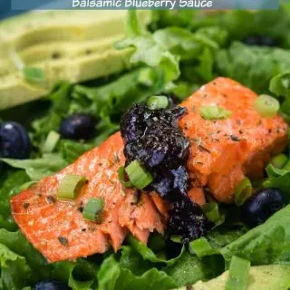 Green lettuce is adorned with a thick piece of salmon topped with a balsamic blueberry sauce. Sliced avocado sits in the background and the recipe title "Salmon Salad with Balsamic Blueberry Sauce" is printed across the top.