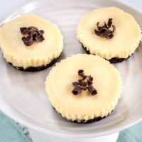Top-down view of 3 Mini Cheesecakes have an Oreo crust, white cheesecake filling and topped with chocolate shavings. They sit on a small cake plate over a blue napkin.