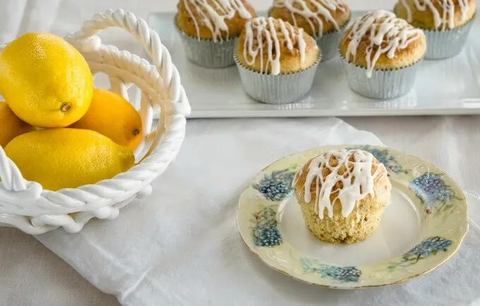Whole lemon pistachio muffin drizzled with a lemon glaze on a floral plate over a white tablecloth. a plate of muffins and a bowl of lemons are sitting in the background.