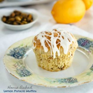 A Lemon Pistachio muffin drizzled with cream cheese glaze sits in the middle of a floral plate over a white tablecloth. A white dish of pistachios and two lemons sit in the background.