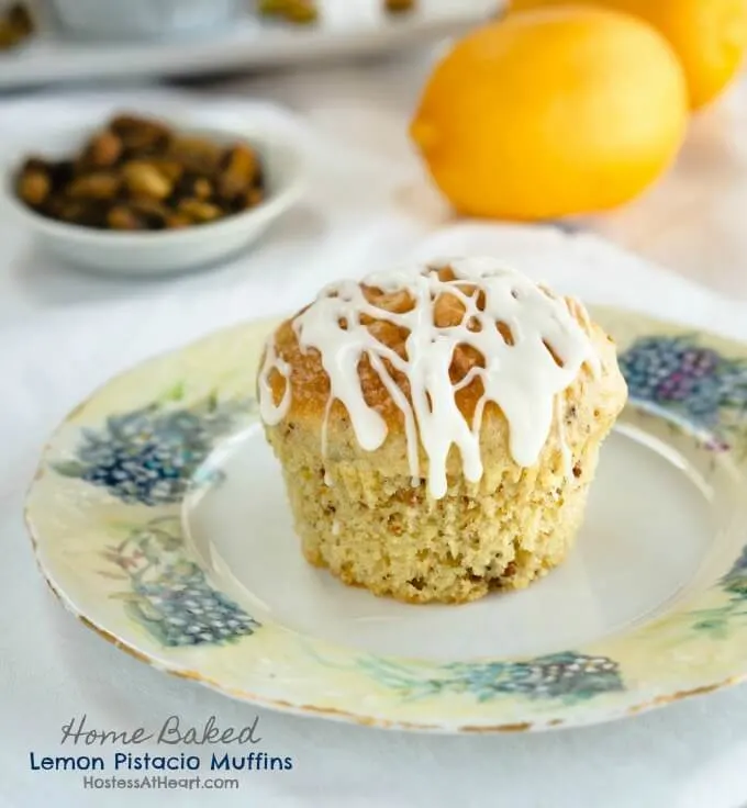 A Lemon Pistachio muffin drizzled with cream cheese glaze sits in the middle of a floral plate over a white tablecloth. A white dish of pistachios and two lemons sit in the background.