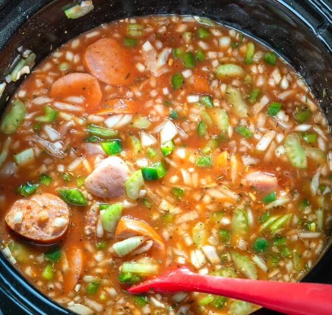 Jambalaya ingredients are added to a slow cooker and stirred with a red spoon.