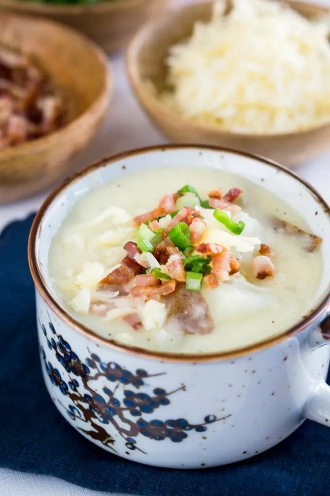 A soup cup filled to the brim with Cheesy Baked Potato Soup topped with more cheese, onion, and bacon. Wooden bowls of bacon and cheese sit in the background.