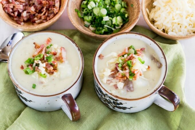 Two soup cups loaded with thick cheesy potato soup topped with cheese, green onion & bacon. Wooden bowls filled with bacon, cheese, and green onions sit in the background.