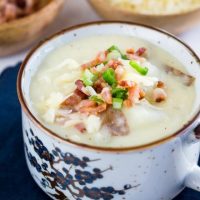 A soup cup filled to the brim with Cheesy Baked Potato Soup topped with more cheese, onion, and bacon. Wooden bowls of bacon and cheese sit in the background.