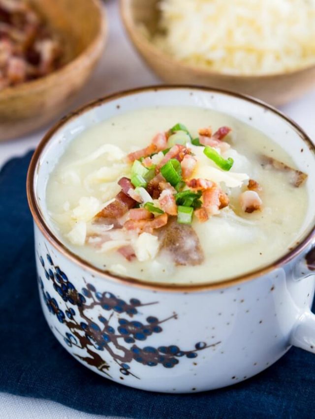 A soup cup filled to the brim with Cheesy Baked Potato Soup topped with more cheese, onion, and bacon. Wooden bowls of bacon and cheese sit in the background.