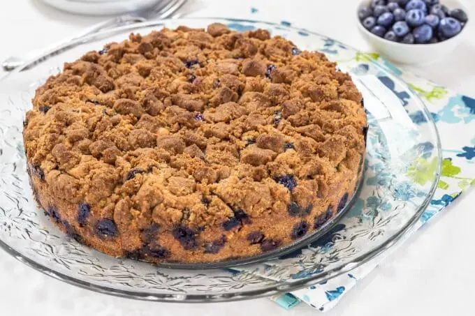 A 9\" round blueberry buckle cake with a thick browned sugar streusel dotted with blueberries around the bottom sitting on a crystal glass plate over a floral napkin.