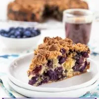 A slice of blueberry buckle showing big berries throughout and topped with an oven-browned streusel topping. The full buckle, a white bowl of blueberries, and a cup of coffee are in the background.