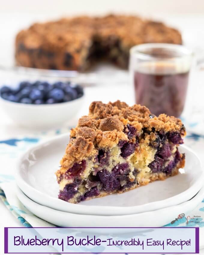 A slice of blueberry buckle showing big berries throughout and topped with an oven-browned streusel topping. The full buckle, a white bowl of blueberries, and a cup of coffee are in the background.