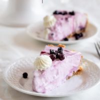 Two slices of Blueberry Lemonade Pie sitting on white plates topped with wild blueberries and a dollop of whipped cream.