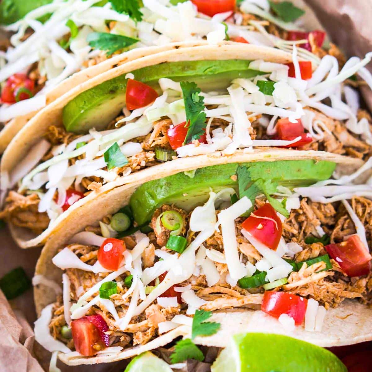 Top down image of 3 shredded chicken tacos garnished with cabbage, cheese, avocado and tomatoes.