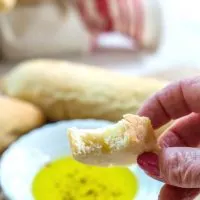 Slice of a Ciabatta Breadstick with a bite take out of it. A white dish filled with olive oil for dipping and more breadsticks it's in the background.