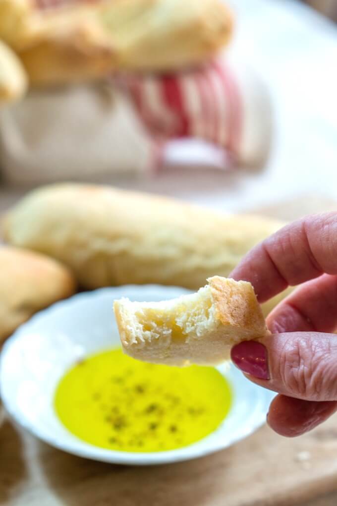 Slice of a Ciabatta Breadstick with a bite take out of it. A white dish filled with olive oil for dipping and more breadsticks it\'s in the background.