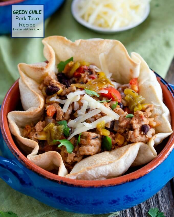 A tortilla bowl sitting in a blue bowl stuffed with ground pork, black beans, corn, and green chilis over a green napkin. The title \"Green Chili Pork Recipe\" appears in the upper left corner.