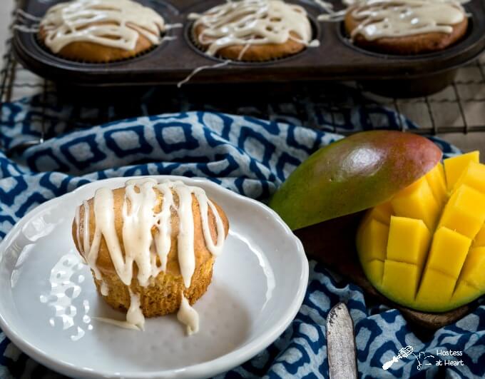 Mango muffin flavored with cream cheese and a vanilla bean drizzle sitting on a plate next to a diced mango over a blue patterned napkin. A full muffin tin sits in the background.