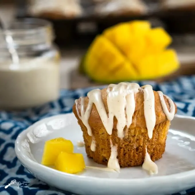 A Cinnamon Ginger Muffin with a drizzle of vanilla bean glaze sits on a white plate over a blue patterned napkin with two pieces of mango. A diced mango sits in the background.