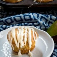 Mango muffin flavored with cream cheese and a vanilla bean drizzle sitting on a plate next to a diced mango over a blue patterned napkin. A full muffin tin sits in the background.