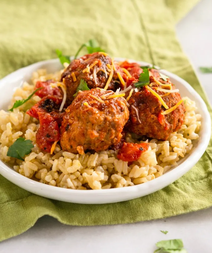 White plate with 4 large Mexican Meatballs covered in Chipotle sauce sitting on a layer of green-tinted tomatillo rice over a green napkin.