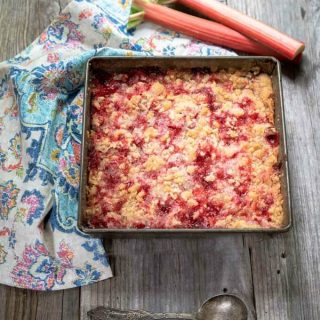 cake pan filled with bright red rhubarb dessert with a crumbled streusel topping surrounded by a multi-colored napkin and fresh rhubarb.