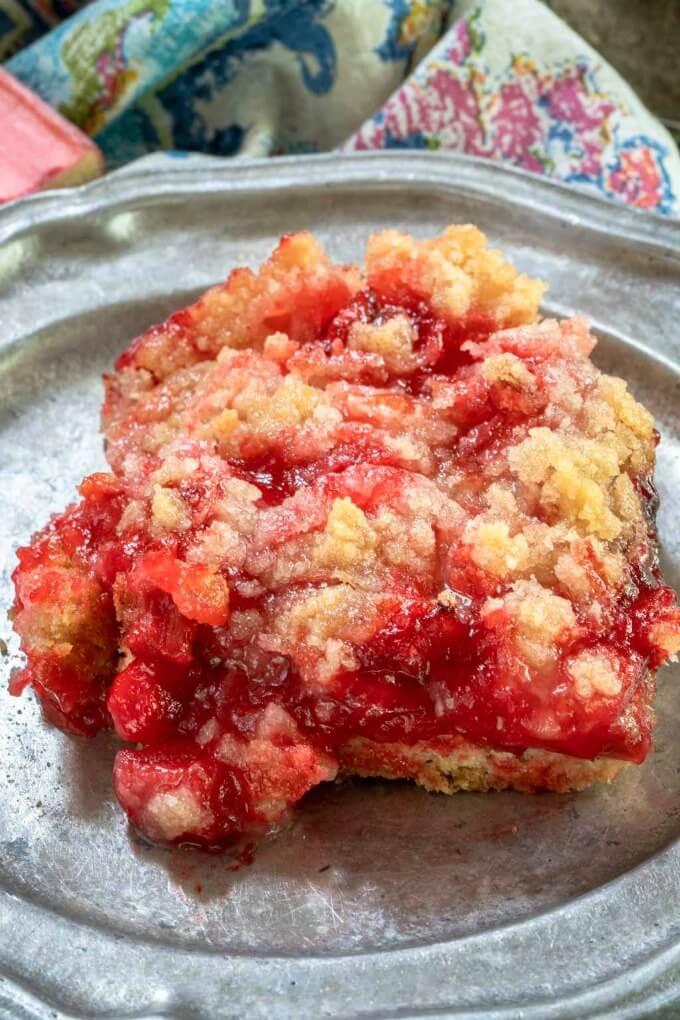 Close up of a bright red Rhubarb dessert bar sitting on a metal plate in front of a multi-color napkin.