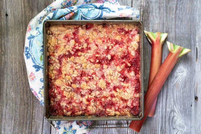 Pan of Rhubarb Cherry Bar Dessert sitting on a barn wood backdrop beside stalks of fresh rhubarb nest to a pretty multi-color napkin.